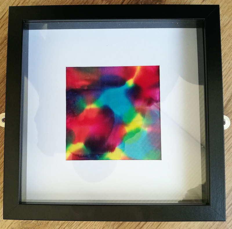 A small square 
 piece of Japanese Silk with abstract shapes and patterns in all the colours of the rainbow. Mounted with a thick border of white card and a deep black square frame