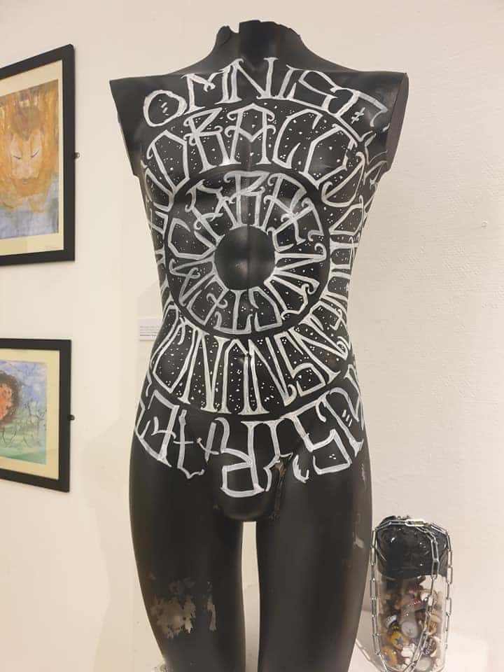 A sculpture of a male body in black plastic  resin with script font painted in white in a circular design